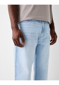 Ljusblå denimjeans med avslappnad passform, som har en något sliten textur och synliga sömnadsdetaljer, ihop med en avslappnad grå topp.