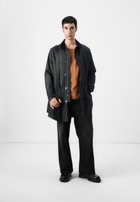 Manteau noir, léger, avec des boutons, doté d'un col à revers. Porté par-dessus une chemise marron et assorti à un pantalon large noir et des chaussures.