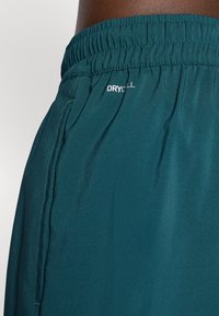 Turkise shorts laget av glatt stoff, med elastisk midjebånd, sidelommer og en liten hvit "DRYCELL"-logo.