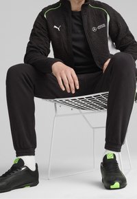 Veste noire matelassée avec des accents verts, t-shirt noir, pantalon de jogging noir et baskets noires avec des détails verts, assis sur une chaise en fil blanc.