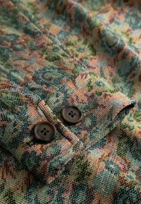 Nahaufnahme einer Manschette aus stoff mit Blumenmuster und zwei dunkelbraunen Knöpfen auf strukturiertem gewebtem Material in Grün-, Orange- und Blautönen.