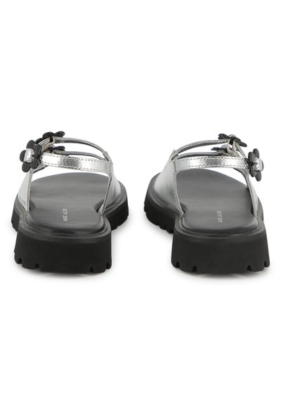 Ασημί μεταλλικά σανδάλια με χοντρή μαύρη ελαστική σόλα, ρυθμιζόμενα λουράκια και διακοσμητικό λουλούδι στο αριστερό λουράκι. Σχεδίαση με επίπεδη σόλα και ανοιχτά δάχτυλα.