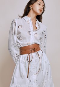 Wit katoenen shirt met geborduurde bloemmotieven, gecombineerd met een tan leren riem die een witte gelaagde rok aandraagt. Knopen zichtbaar.