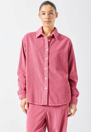 Donna che indossa una camicia larga di velluto a coste rosa con bottoni e pantaloni abbinati, in piedi contro uno sfondo bianco semplice.