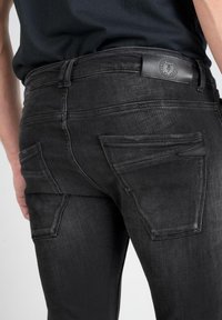 Jeans en denim noir avec un design ajusté, taille mi-haute, et deux poches arrières à patch avec une finition texturée. Patch en cuir avec logo à la taille.