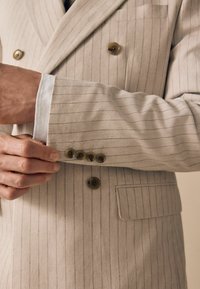 Beige double-breasted suit jacket with light stripes. Features gold-tone buttons and a white shirt cuff. Texture appears smooth and tailored.