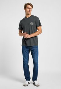 Camiseta de manga corta gris oscuro con un pequeño logo impreso en blanco en el pecho, combinada con jeans de mezclilla azules y zapatillas blancas.