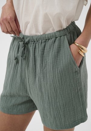 Personne portant un short vert texturé avec une taille élastique et un cordon, main dans la poche, et un bracelet en or au poignet.