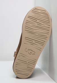 Tan suede saapas, jossa on teksturoitu kevyt pohja, jossa on huomattava UGG-logo ja yksityiskohtainen urapinta pitoa varten.