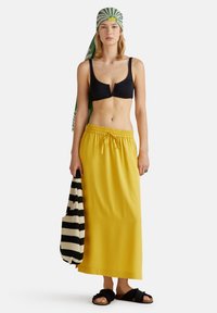 Zwarte bikini-top, gele maxi-rok, groen en wit gestreepte hoofddoek, zwart-wit gestreepte tas, en zwarte fuzzy sandalen.