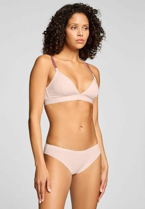 Frau mit lockigem Haar, die einen hellrosa BH und passende Unterwäsche trägt, steht vor einem schlichten hellgrauen Hintergrund.