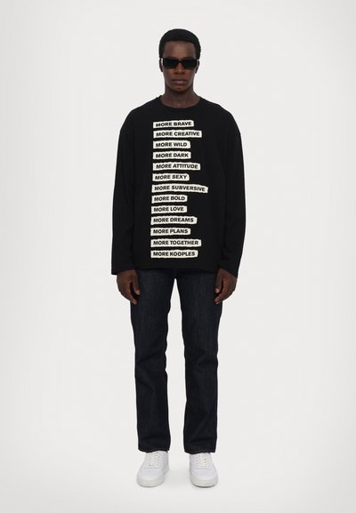 Black sweatshirt with white text phrases on the front, paired with dark jeans and white sneakers. Model wearing sunglasses.
