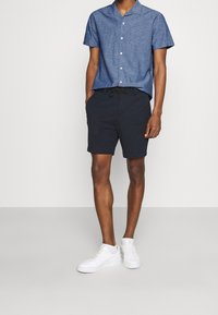 Camicia a maniche corte con bottoni in tessuto chambray blu abbinata a pantaloni corti blu navy e sneakers bianche. Fit casual e rilassato.