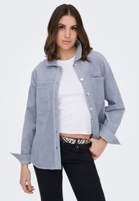 Noorpruunide õlgadeni juustega noor naine, kes kannab hele sinist korduroost jopet valge lühikese topi ja mustade teksade peal.