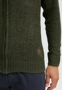 Zelený žebrovaný pletený kardigan s texturovaným vzorem a koženým štítkem. Má přední zip a design stahovacího manžetu.