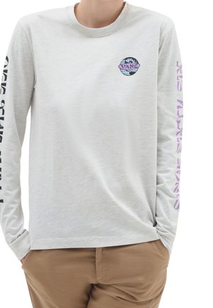 Osoba ubrana w jasnoszarą koszulę z długim rękawem z logo globu na piersi i tekstem na obu rękawach, w połączeniu z brązowymi spodniami.