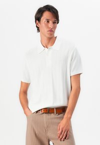 Man standing with hands in pockets wearing a white short-sleeve polo shirt, brown belt, and beige pinstripe trousers on white background.