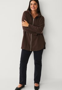 Brown zip-up hoodie with a hood, soft texture, long sleeves, side slits, paired with dark trousers and black flat shoes.