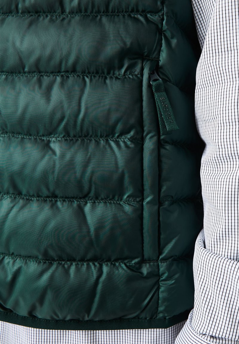 Chaleco acolchado en verde oscuro con costuras horizontales. Presenta un bolsillo con cremallera y un acabado texturizado y suave. Se lleva sobre una camisa de cuadros.