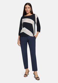 Top van geborstelde stof met geometrische patronen in marineblauw en crème, driekwart mouwen, gecombineerd met marineblauwe broek en zwarte sandalen met bandjes.