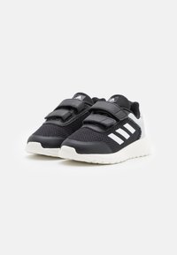 Baskets pour enfants Adidas en noir et blanc, en tissu en maille, avec des bandes velcro et des semelles blanches sur un fond clair.