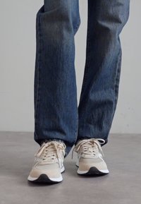 Beige og grå treningssko laget av mesh og semsket skinn, med runde snørebånd og svart såle. Brukt med mørkeblå jeans med rett ben.