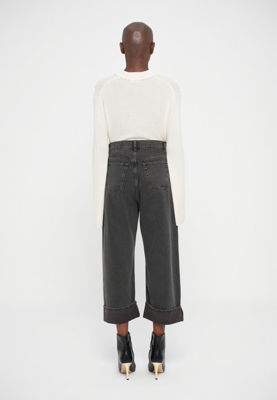 Person som har på seg en hvit strikkegenser, høy midje vid benkorte svarte jeans og svarte hæl ankelstøvler, står vendt bort.