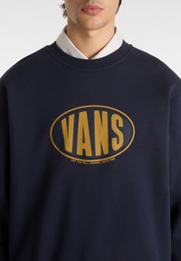 Sudadera de color azul marino hecha de algodón, con un gran logo ovalado dorado que presenta el texto "VANS" y un eslogan más pequeño debajo. Escote redondo.
