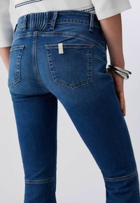 Personne portant un jean en denim bleu avec des poches arrière et un haut blanc, avec des bracelets argentés au poignet droit, sur un fond uni.