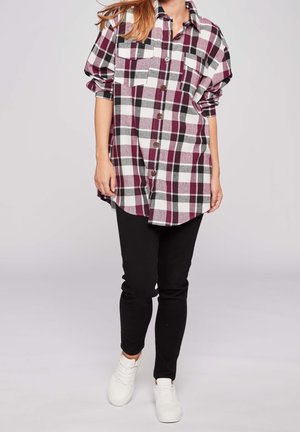 Persoon die een lange geruite blouse in bordeauxrood, zwart en wit draagt, zwarte broek en witte sneakers, staand tegen een effen achtergrond.