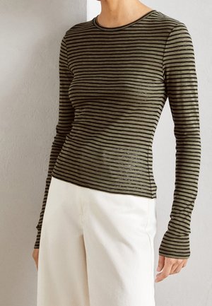 Personne portant un haut ajusté à manches longues rayé vert olive et noir avec un pantalon blanc taille haute, debout contre un mur clair.