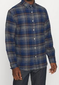 Blå och grå rutiga flanellskjorta med bruna accenter, knapp-upp design och långa ärmar. Har en klassisk krage och texturerad tyg.