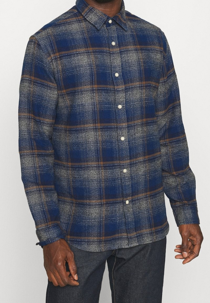 Blå och grå rutiga flanellskjorta med bruna accenter, knapp-upp design och långa ärmar. Har en klassisk krage och texturerad tyg.