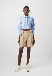 Cropped blue striped shirt with long sleeves and beige pleated shorts. White sneakers complete the outfit. Neutral background.
