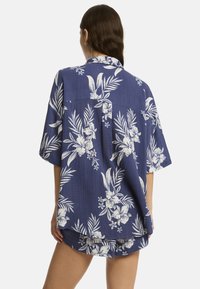 Modelo vistiendo una camisa floral de corte holgado en azul marino con pantalones cortos a juego. La camisa presenta flores y hojas tropicales en color blanco.