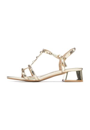 Sandalias de tacón metálicas doradas con diseño de tiras, tacón bajo en bloque y detalles con tachuelas. Construcción con punta abierta, forma elegante y moderna.