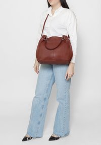 Sac à main en cuir marron avec deux poignées courtes, une bandoulière amovible et des accessoires en argent. Le sac présente une forme arrondie et une texture lisse.