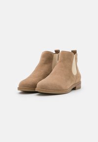 Bottines en daim marron avec des panneaux élastiques sur les côtés, bout arrondi et un petit talon empilé. Elles présentent une texture lisse et un design simple.
