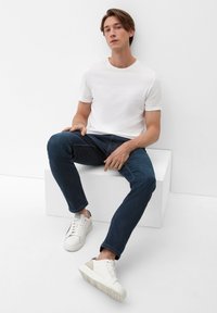 Witte crewneck t-shirt, donkerblauwe slim-fit jeans en witte sneakers met grijze en beige accenten. Zittend op een witte kubus tegen een effen achtergrond.