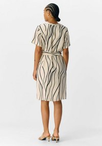 Beige jurk met korte mouwen en zwart golflijnenpatroon, met een taillering en een lengtes tot de knie. Model draagt neutrale open teen hakken.
