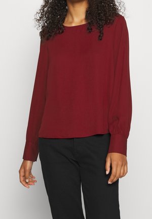 Kobieta w bordowej bluzce z długim rękawem z mankietami i czarnych spodniach, stojąca na tle jasnego, jednolitego tła.