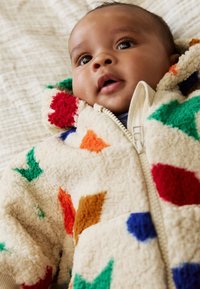 Veste en polaire couleur crème avec une fermeture éclair, ornée de patchs géométriques colorés en rouge, vert, bleu et orange. Texture épaisse et design à capuche.