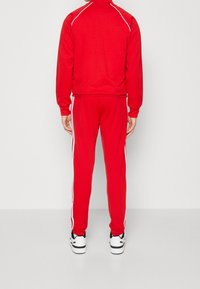 Röd träningsoverall med vita detaljer, zip-up jacka och byxor med elastisk midja. Tillverkad av ett mjukt, lätt material. Atletisk passform.