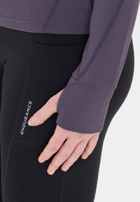 Pitkähiha, kapeamuotoinen paita tummasta violetista kankaasta, joka on yhdistetty mustiin leggingseihin, joissa on sivutasku ja valkoinen "ENDURANCE"-logo. Sileä pinta.