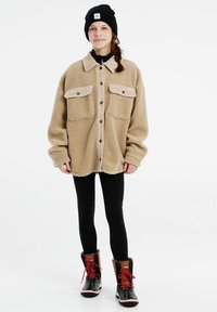 Veste en laine polaire beige avec fermeture à boutons, deux poches avant et col côtelé. Portée sur des leggings noirs et des bottes d'hiver avec des lacets rouges.