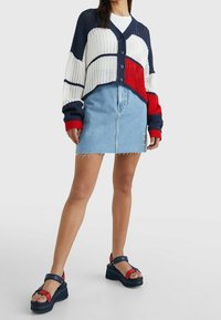 Cardigan lavorato a maglia rosso, bianco e blu navy con design corto e bottoni, abbinato a una gonna di jeans azzurro chiaro con dettagli a frange. Sandali con zeppa.