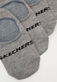 Három pár szürke, láthatatlan zokni, kék sarokpárnával és fekete Skechers logóval a lábujjaknál, egy fehér felületen, átfedésben elhelyezve.