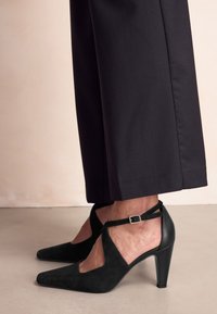 Czarne szpilki z noskiem w szpic, wykonane z zamszu i skóry. Posiadają pasek wokół kostki z złotą klamrą oraz wycięcia w designie.