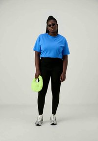 Woman wearing blue t-shirt, black leggings, silver sneakers, sunglasses, holding neon green fluffy handbag, standing against plain background.