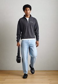 Mørkegrå lynlås sweatshirt, hvid t-shirt, lyseblå jeans, sorte loafers, bærende en lille sort taske, stående på trægulv.